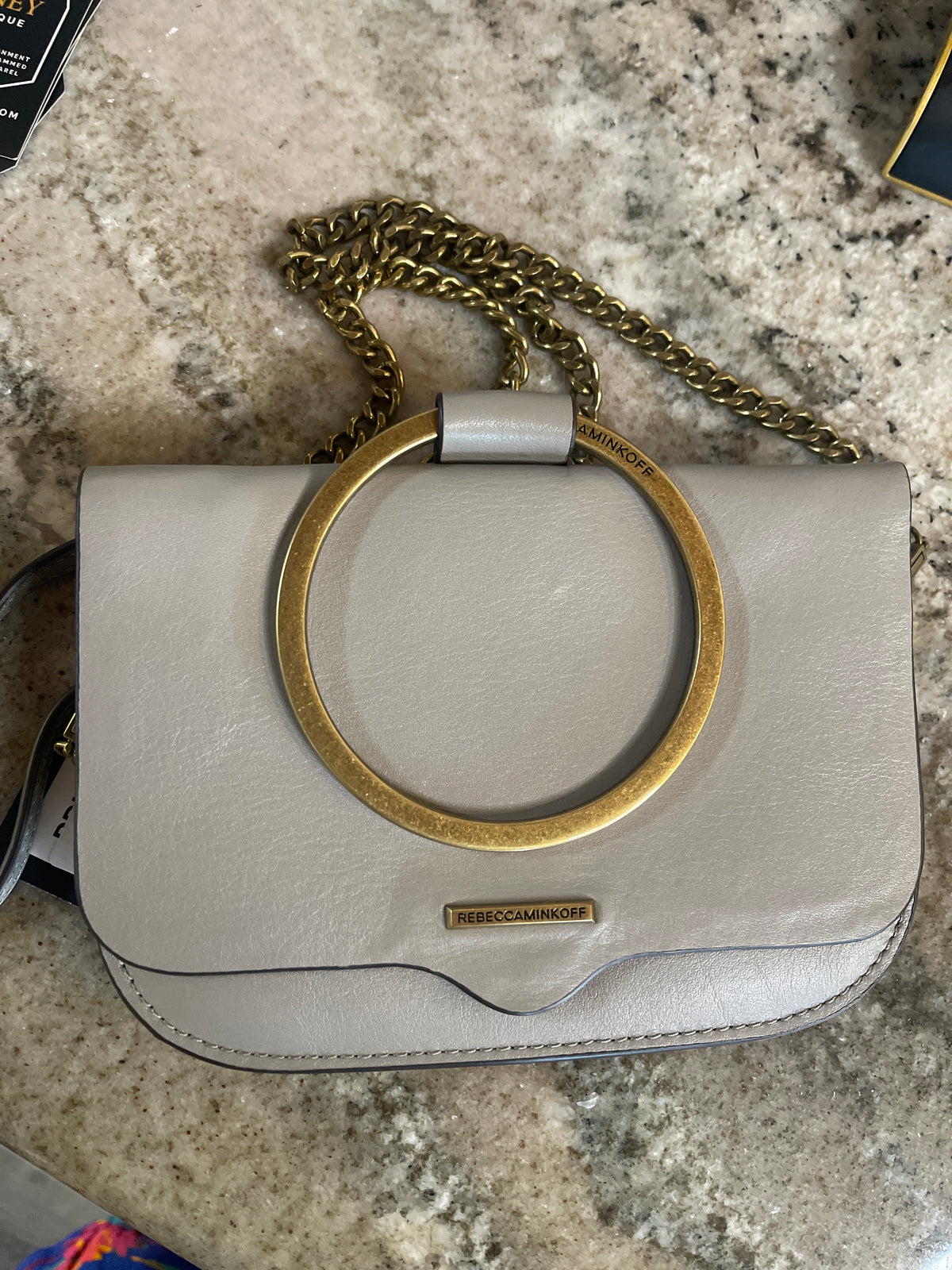 The handbag pictured is the Rebecca Minkoff Ring Crossbody (often referred to as the Ring Clutch or Wristlet) in a taupe or light grey leather with antiqued gold-hardware and top ring handle.