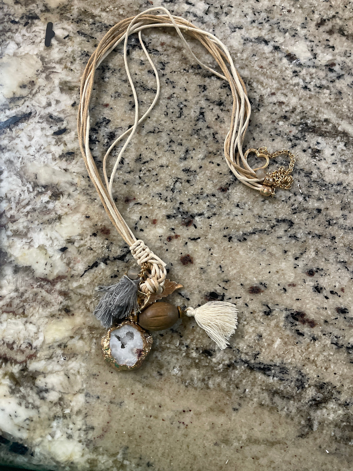 Bohemian-style charm necklace or decorative tassel pendant featuring multi-strand cords, a white tassel, a faux-agate slice with gold edging, a wooden bead, and a gold-tone leaf charm.